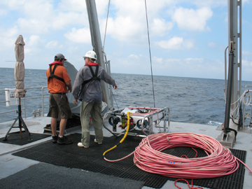 Two men standing near an ROV and its coiled umbilical on the back deck of a boat.