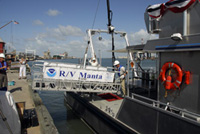 A small crane mounted on the back deck of the Manta is used to swing the gangway into place between the boat the the pier.  A banner with the NOAA logo and R/V Manta on it hangs from the side of the gangway.