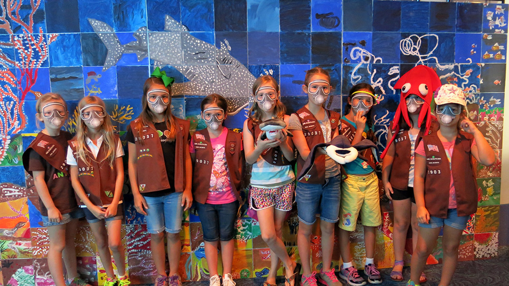 A group of girls in Brownie uniforms wearing paper scuba masks and standing in front of a coral reef mural