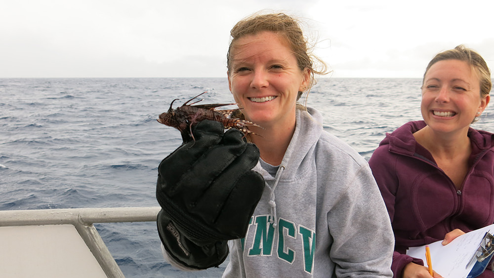 Two women on a boat--one holding up a small lionfish, the other with a clipboard to record data