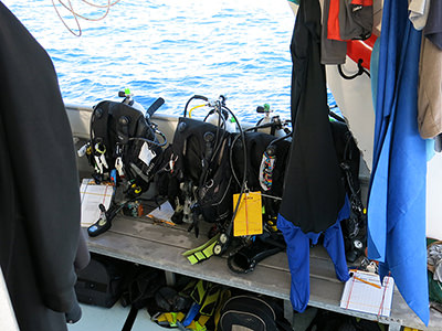 Scuba gear lined up on a dive bench on a boat with wetsuits hanging nearby