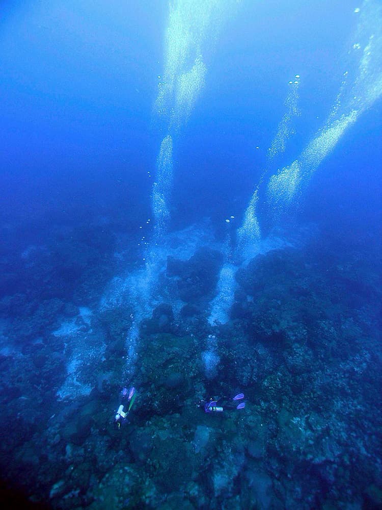 Two divers on the reef far below with their bubbles streaming upward.